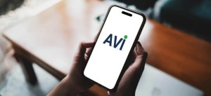 Pessoa segurando smartphone com logo da AVA na tela, em ambiente interno com mesa de madeira ao fundo, representando tecnologia e inovação.