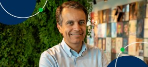 Homem sorridente de cabelo castanho claro e camisa social azul claro, posando em ambiente com parede de livros e parede de plantas verdes ao fundo.