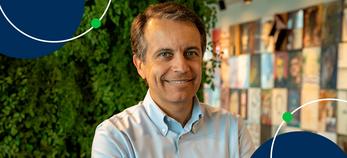 Homem sorridente de cabelo castanho claro e camisa social azul claro, posando em ambiente com parede de livros e parede de plantas verdes ao fundo.