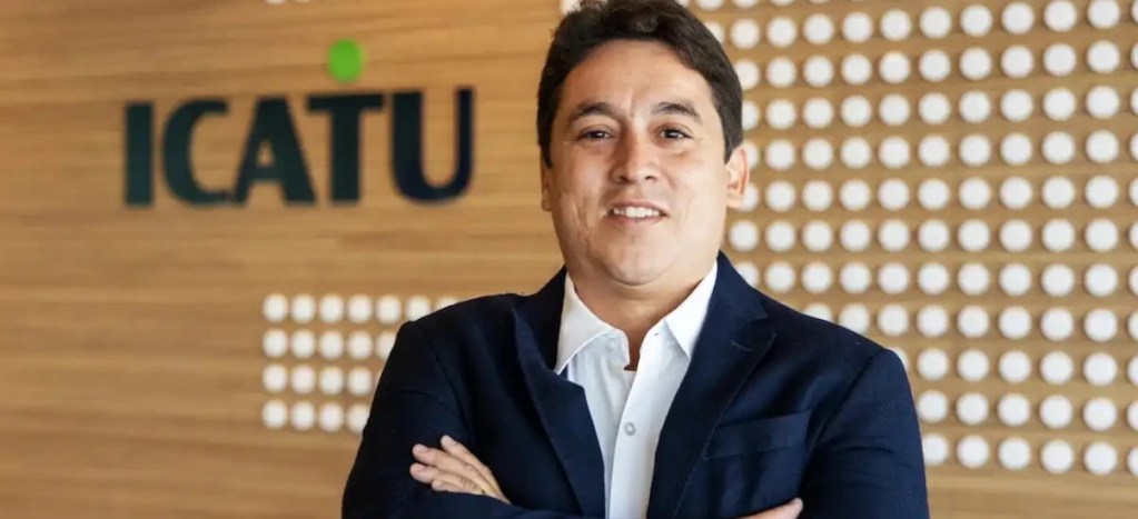 Homem sorridente vestindo terno azul escuro e camisa branca, com os braços cruzados na frente de um fundo de madeira com o logo ICATU, representando um profissional de seguros ou corretor de seguros.