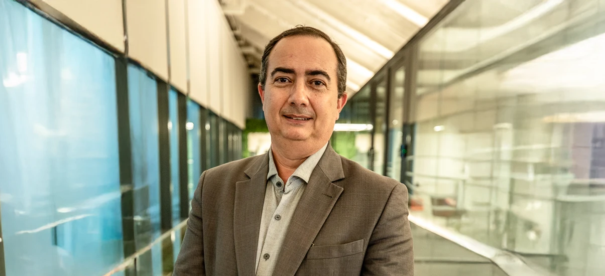 Homem profissional em ambiente moderno, usando terno e camisa cinza claro, sorrindo para a câmera em um corredor de vidro e metal.