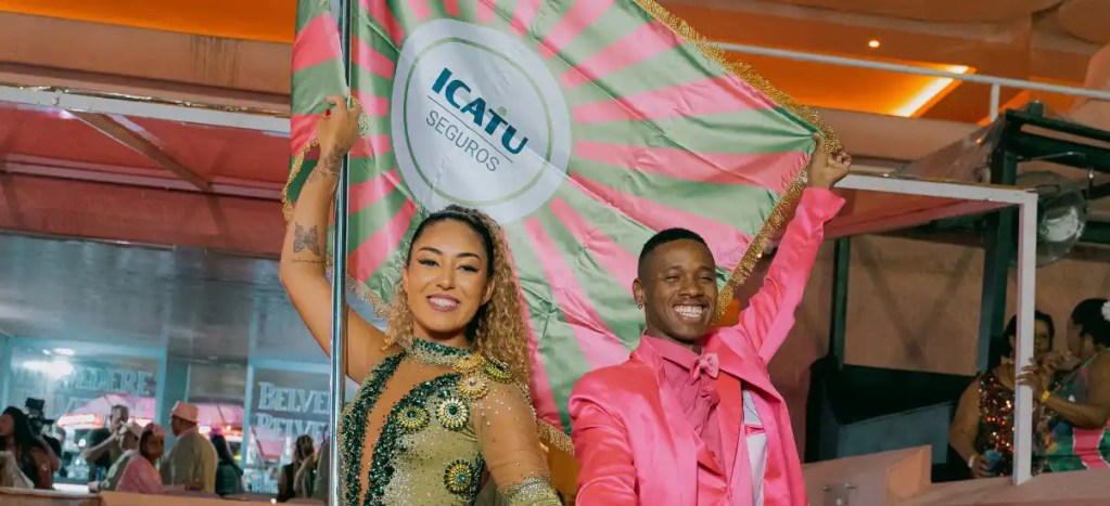 Dançarinos comemorando com bandeira da ICATU Seguros em evento festivo, vestindo roupas brilhantes e sorrindo, transmitindo alegria e celebração.