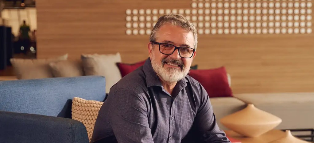 Homem sorridente com barba grisalha e óculos de grau sentado em um ambiente acolhedor com sofá e decoração moderna ao fundo